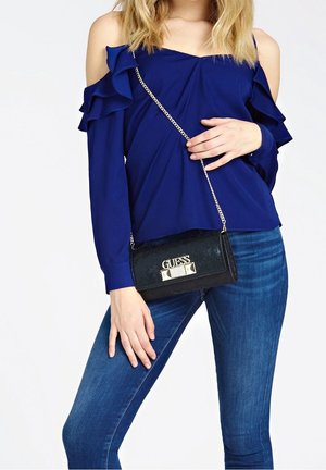 Marineblauwe off-shoulder blouse met geruite mouwen, gecombineerd met een zwarte faux-lederen crossbodytas met een zilveren logo en kettingriem.