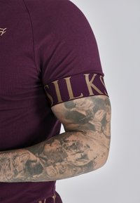 Fioletowa koszula z krótkim rękawem o dopasowanym kroju, z złotą tasiemką "SILKS" wokół rękawa. Ramię zdobią misternie wykonane tatuaże.