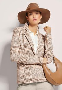 Gepatineerde bruine en crème geblokte blazer met een gekleurde kraag, gedragen over een witte blouse, gecombineerd met een bruine leren tas en een brede hoed.