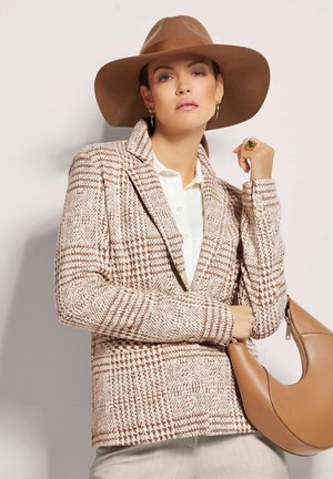Gepatineerde bruine en crème geblokte blazer met een gekleurde kraag, gedragen over een witte blouse, gecombineerd met een bruine leren tas en een brede hoed.