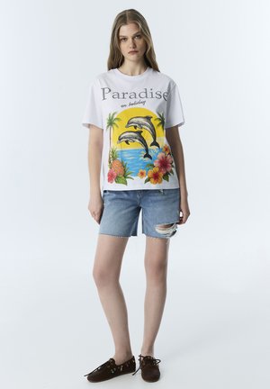 Junge Frau trägt ein weißes T-Shirt mit Delfinen, tropischen Blumen und dem Schriftzug „Paradise on holiday“, kombiniert mit zerrissenen Jeansshorts und braunen Schuhen.