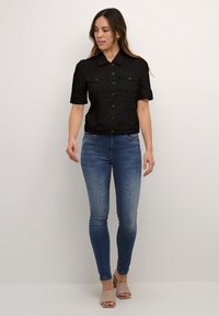 Svart croppad jacka med knappar framtill och korta ärmar, med två bröstfickor. Bärs över blå skinny jeans och beige öppna klackar.