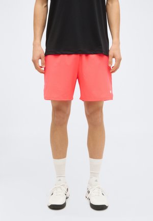 Homme portant un short de sport corail vif, un haut noir, des chaussettes blanches courtes et des baskets blanches, debout sur un fond clair uni.