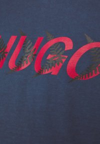 T-shirt in cotone blu navy con il testo "HUGO" rosso intenso e dettagli in felce verde scuro. Texture morbida, design casual, ripresa ravvicinata.