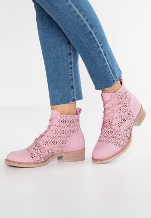Femme portant des bottines lacées rose clair avec des motifs floraux découpés au laser et un jean court bleu clair sur fond blanc.