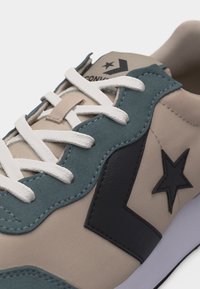 Κοντινό πλάνο μπεζ και πετρόλ αθλητικού παπουτσιού με λευκούς κορδόνια και μαύρο αστέρι και λοξό σήμα στη πλευρά.