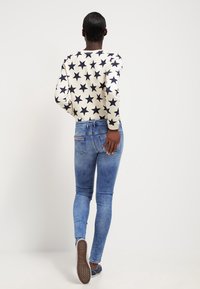 Langärmliges cremefarbenes Oberteil mit navyblauem Sternenprint, kombiniert mit engen blauen Jeans. Die Rückansicht zeigt eine glatte Textur und einen lässigen Schnitt.