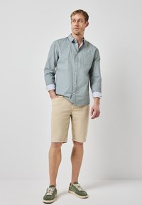 Lehce pruhovaná zelená košile s límečkem, béžové šortky a zelené tenisky s bílými tkaničkami, stojící na jednobarevném pozadí.