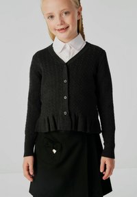 Cardigan lavorato a maglia scuro con scollatura a V, texture a coste, cinque bottoni e orlo arricciato, indossato sopra una camicia bianca con colletto e una gonna nera.