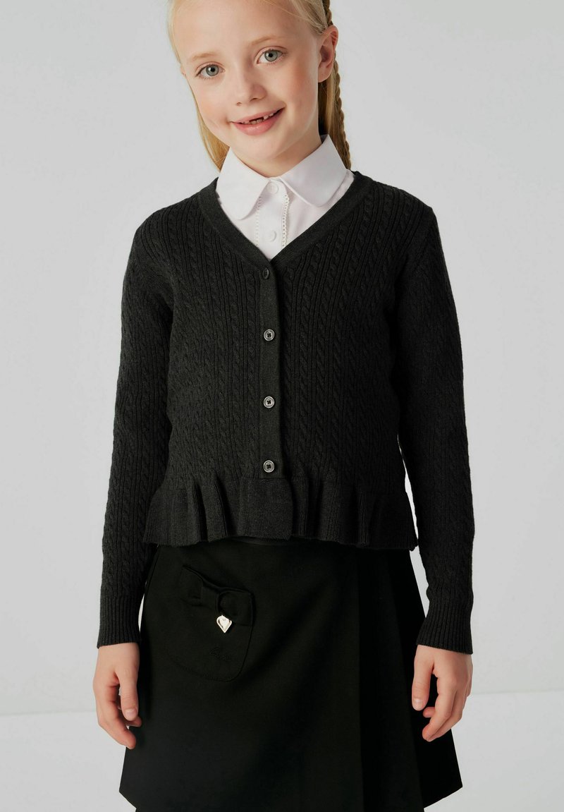 Cardigan lavorato a maglia scuro con scollatura a V, texture a coste, cinque bottoni e orlo arricciato, indossato sopra una camicia bianca con colletto e una gonna nera.