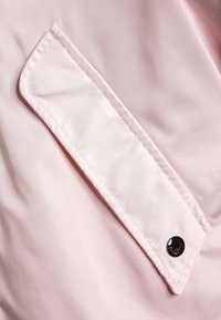 Ljusrosa tyg med en slät textur och sydda detaljer. Har en rektangulär klaff med en svart tryckknapp.