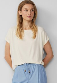 Lichtbeige T-shirt met korte mouwen en een wijde halslijn, gecombineerd met lichtblauwe broek met trekkoord en decoratieve kraalaccenten.