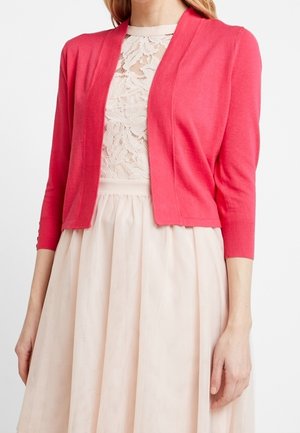 Femme portant un cardigan corail rose sur un haut en dentelle blanche et une jupe plissée rose clair, vue du cou à la taille.