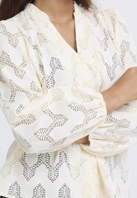 Cream blouse with textured pattern, featuring a geometric lace design. Gathered sleeves and button details enhance its structure.