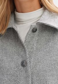 Gros plan sur une personne portant un col roulé blanc et un manteau gris à rayures avec deux boutons noirs visibles attachés.