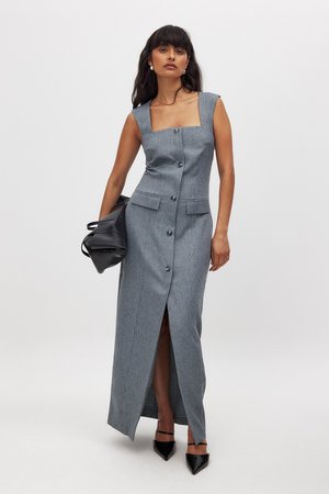 Robe maxi grise sans manches avec décolleté carré, boutons sur le devant, poches à rabat et fente sur le côté. Associée à un sac à main noir et des escarpins.
