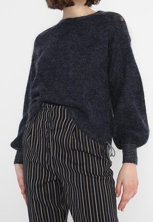 Kvinde iført en mørk, blød sweater med ballonsærmer og stribede bukser med høj talje og knapper foran, stående mod en hvid baggrund.