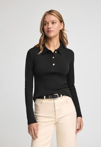 Polo shirt nero a maniche lunghe con colletto e quattro bottoni dorati, abbinato a jeans beige chiaro a vita alta e una cintura nera.