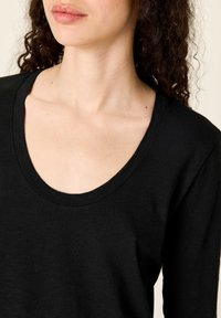 Naine, kellel on lokkis tume juuksed ja musta kaelusega pika varrukaga särk, kujutatud õlgadest kuni huulteni heleda tausta ees.