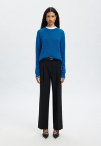 Spilgti zils, teksturēts džemperis apvienots ar melniem, platajiem biksēm. Modelis valkā melnas, norādītes apavus un tievu melnu jostu. Zem džempera ir vienkārša balta krekla.