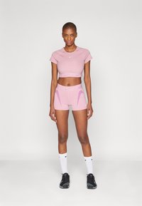 Lyserosa treningscrop-top og shorts med lilla detaljer, laget av glatt stoff. Modellen har på seg svarte joggesko og hvite ankelsokker.