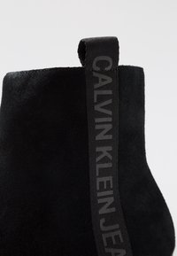 Musta mokkanahkainen nilkkaboots, jossa on teksturoitu pinta ja silmiinpistävä musta logo-nyöri helppoa pukemista varten. Sujuva siluetti, jossa on minimaalinen muotoilu.