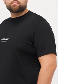 Schwarzes Baumwoll-T-Shirt mit Rundhalsausschnitt, kurzen Ärmeln und einem weißen "JJ Studios"-Logo auf der Brust. Glatte Textur, reguläre Passform.
