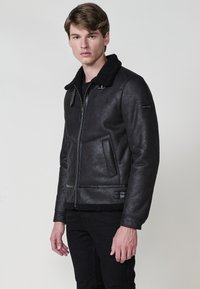 Giacca in ecopelle nera con finitura testurizzata, colletto in shearling, chiusura con zip, tasche laterali e branding discreto sulla manica.