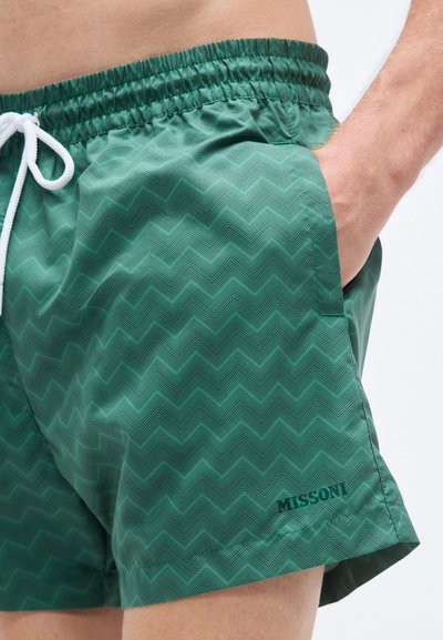 Πράσινα μαγιό σορτς με ζιγκ-ζαγκ μοτίβο, κατασκευασμένα από ελαφρύ υλικό. Διαθέτουν ελαστική μέση με κορδόνι και πλαϊνή τσέπη.