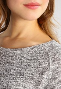 Grå och vit stickad tröja med texturerad yta, bred halsringning och synliga sömmar längs halskanten. Närbild på halsringning och tyg.