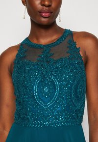 Rochie din mărgele teal cu un guler din plasă transparentă, broderie florală intricate și modele texturate detaliate. Bust ajustat care se continuă cu o fustă fluidă.