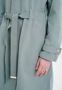 Light grey, belted coat in a smooth, lightweight fabric with two side pockets. Features a buttoned cuff and a decorative silver hardware accent.