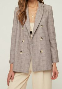 Blazer doppiopetto grigio a quadri con grandi bottoni, rever a lancia e vestibilità sartoriale. Tessuto morbido e forma strutturata con tasche laterali.