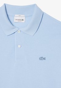 Ljusblå polo skjorta med texturerad tyg, lössittande passform, två-knapps knäppning och broderad Lacoste krokodil-logga på vänster bröst.