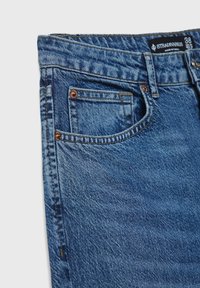 Jeans in denim di blu medio con un classico design a cinque tasche, caratterizzati da rivetti e dettagli di cucitura in rame, che si restringono verso la gamba.