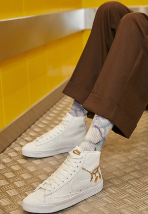 Baskets montantes blanches avec une semelle texturée, détaillées d'un accent de chaîne dorée, associées à des chaussettes tie-dye multicolores et un pantalon large marron.