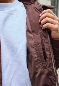 Brun, lynlåsjakke med en glat, blank overflade og en indvendig lomme; lagd over en lyseblå sweatshirt.