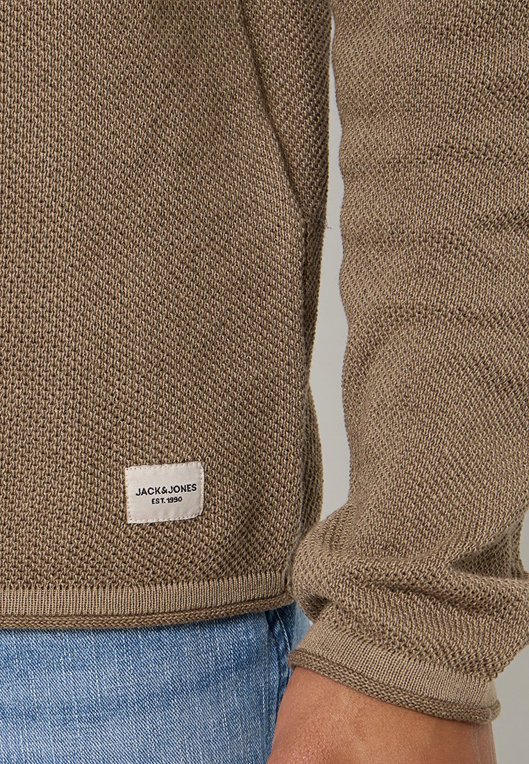 Brauner gestrickter Pullover mit gerippten Bündchen und Saum. Mit einem weißen Etikett mit "JACK & JONES EST. 1990" auf der linken unteren Seite. Sichtbare blaue Jeans.