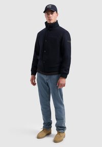 Marineblauwe wollen blendjas met een hoge kraag, knoopsluiting, ribgebreide manchetten en schouderpatch. Gecombineerd met lichte denim jeans en tan kleurige laarzen.