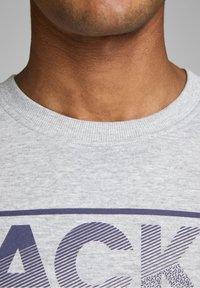 Felpa grigia in cotone con girocollo, caratterizzata da una stampa grafica in blu scuro. Il tessuto ha una consistenza liscia con dettagli a costine sul collo.