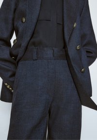 Blazer bleu marine ajusté avec des boutons dorés, associé à un pantalon taille haute à jambes larges assorti et un chemisier noir.