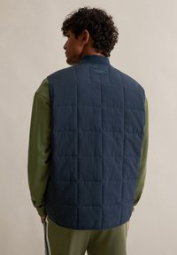 Marineblaue gesteppte Weste mit Stehkragen, versehen mit einem dezenten Logotag auf der Rückseite. Kombiniert mit einem grünen Langarmshirt darunter.