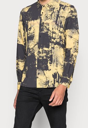 Mann trägt ein langärmeliges Hemd mit Knöpfen und einem abstrakten gelb-schwarzen Sprenkelmuster, kombiniert mit schwarzer Hose.