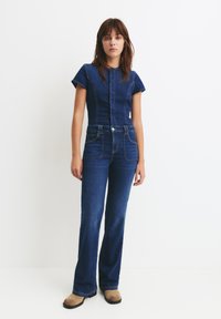Dżinsowy kombinezon w głębokim niebieskim kolorze z krótkimi rękawami, dopasowaną górą i rozszerzanymi nogawkami. W połączeniu z beżowymi botkami za kostkę.