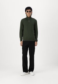 Pull tricot zippé vert avec des poignets côtelés, associé à un pantalon cargo noir et des baskets blanches. Le modèle porte des lunettes de soleil rondes.