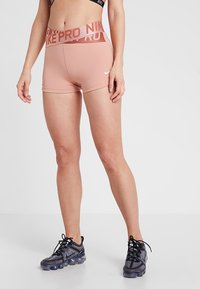 Žena v růžových šortkách Nike Pro a černých teniskách Nike stojí proti jednoduchému světlému pozadí.