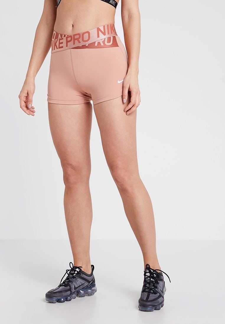 Žena v růžových šortkách Nike Pro a černých teniskách Nike stojí proti jednoduchému světlému pozadí.