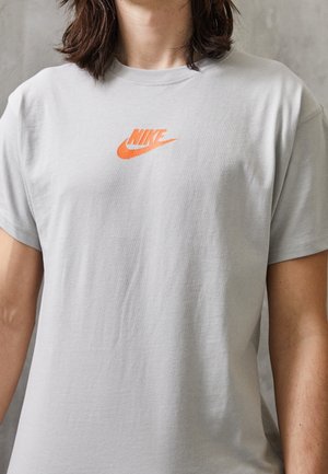 Jasnoszara koszulka z krótkim rękawem, wykonana z bawełny, z pomarańczowym logo Nike umieszczonym na środku klatki piersiowej. Gładka tekstura, standardowy krój.