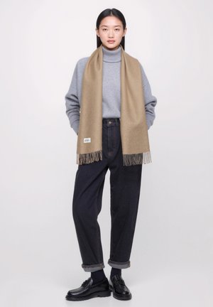 Écharpe en laine beige avec des franges, portée avec un pull à col roulé gris et un jean en denim foncé. Des chaussures noires complètent la tenue.