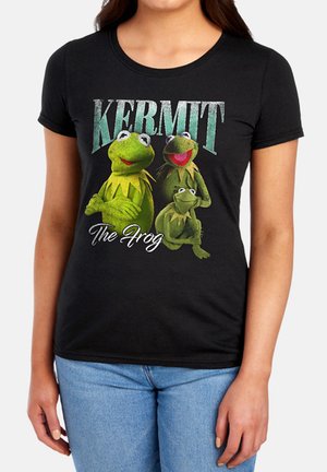Frau trägt ein schwarzes T-Shirt mit drei Kermit der Frosch Figuren und dem Text "KERMIT The Frog" auf der Vorderseite.